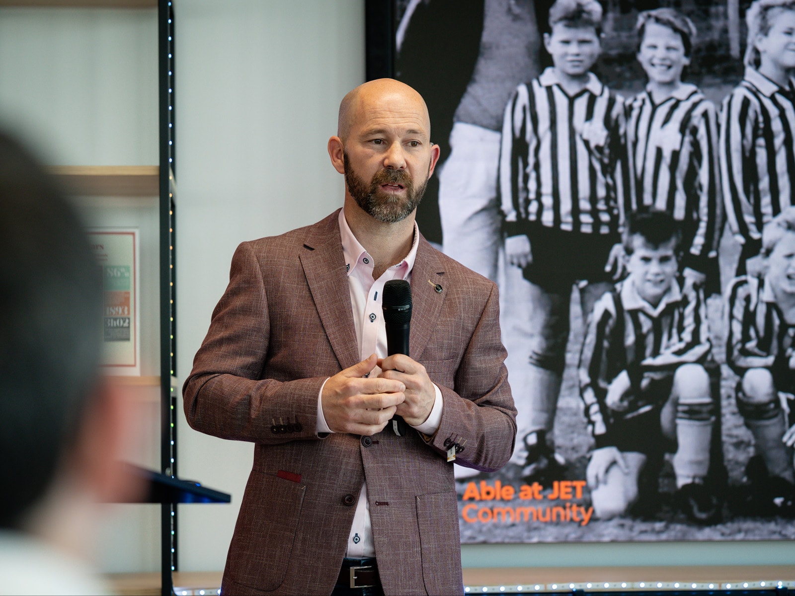 Carl Morris speaking at a microphone, a black and white school football team photo on the screen behind him